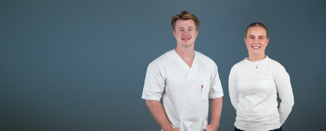 To studenter se ri kamera og smiler. En av dem har på seg sykepleieruniform.