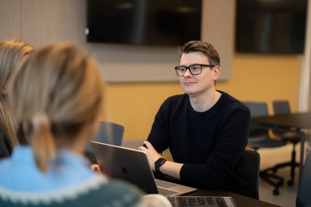 Ung mann sitter i et møterom sammen med andre
