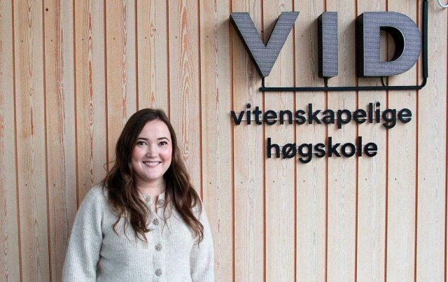 Ung dame med brunt hår poserer foran en vegg med VID-logoen i bakgrunnen