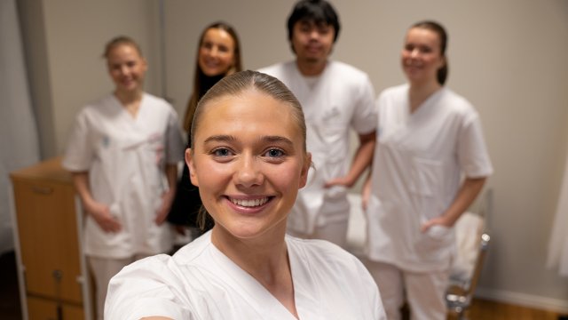 Selfie av jente i sykepleieruniform. I bakgrunnen er flere studenter.
